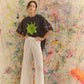 Person wearing a black t-shirt with a green design and white pants standing against a colorful abstract wall.