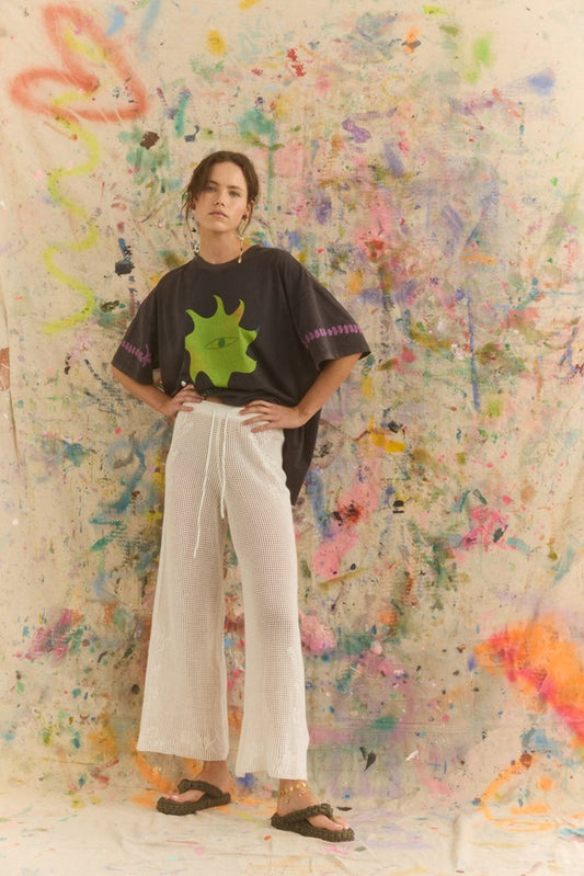 Person wearing a black t-shirt with a green design and white pants standing against a colorful abstract wall.