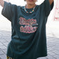 Woman wearing a dark green t-shirt with colorful text and patterned pants on a street.