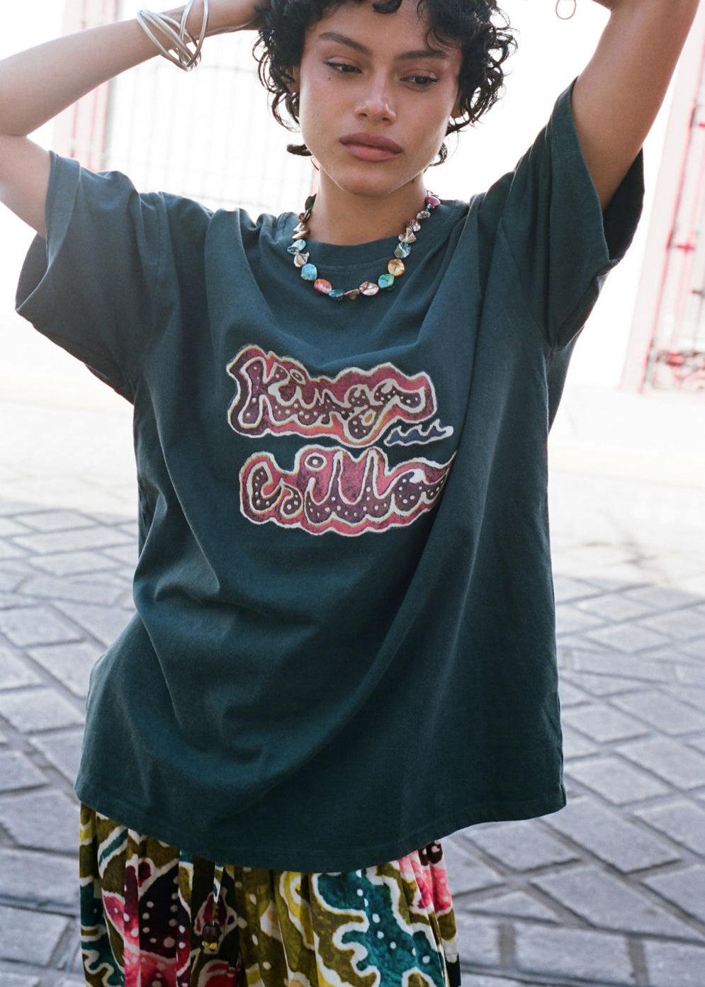 Woman wearing a dark green t-shirt with colorful text and patterned pants on a street.
