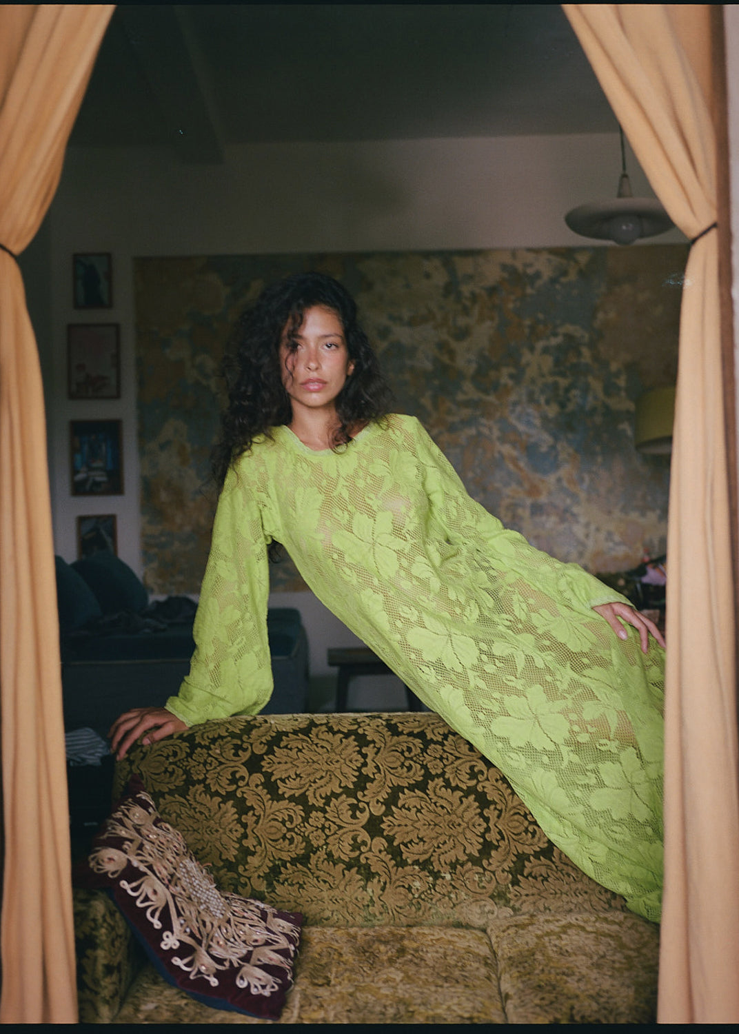 Woman in a green dress standing in a room with patterned walls and curtains.