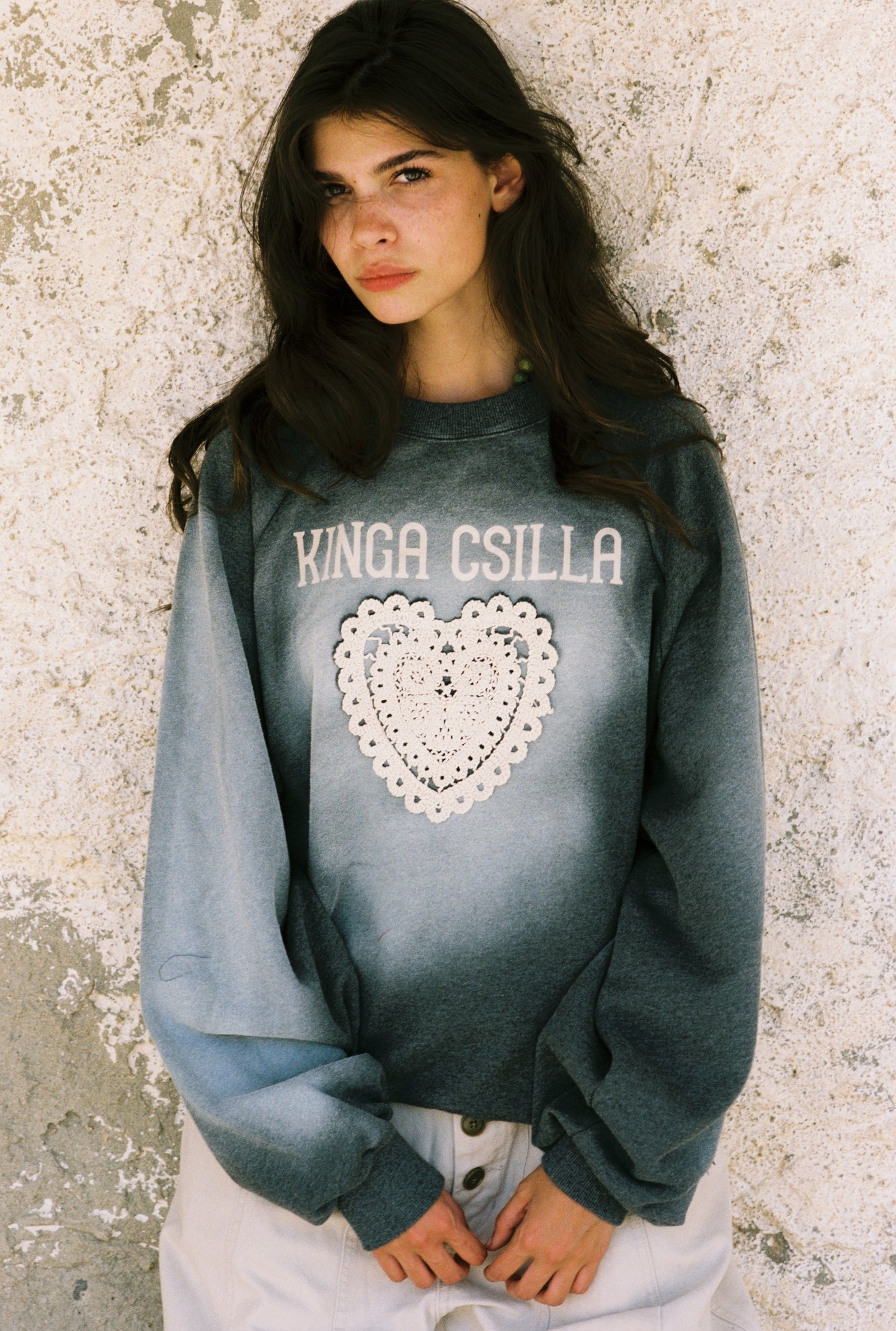 Woman wearing a gray sweatshirt with 'KINGA CSILLA' and a heart design against a textured wall.