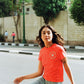 Woman in a red t-shirt and jeans standing on a street with trees and buildings in the background