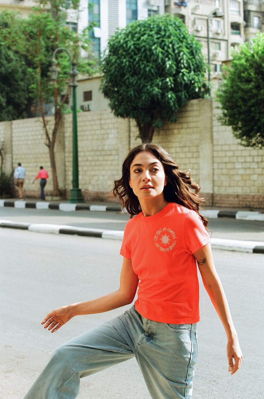 Woman in a red t-shirt and jeans standing on a street with trees and buildings in the background