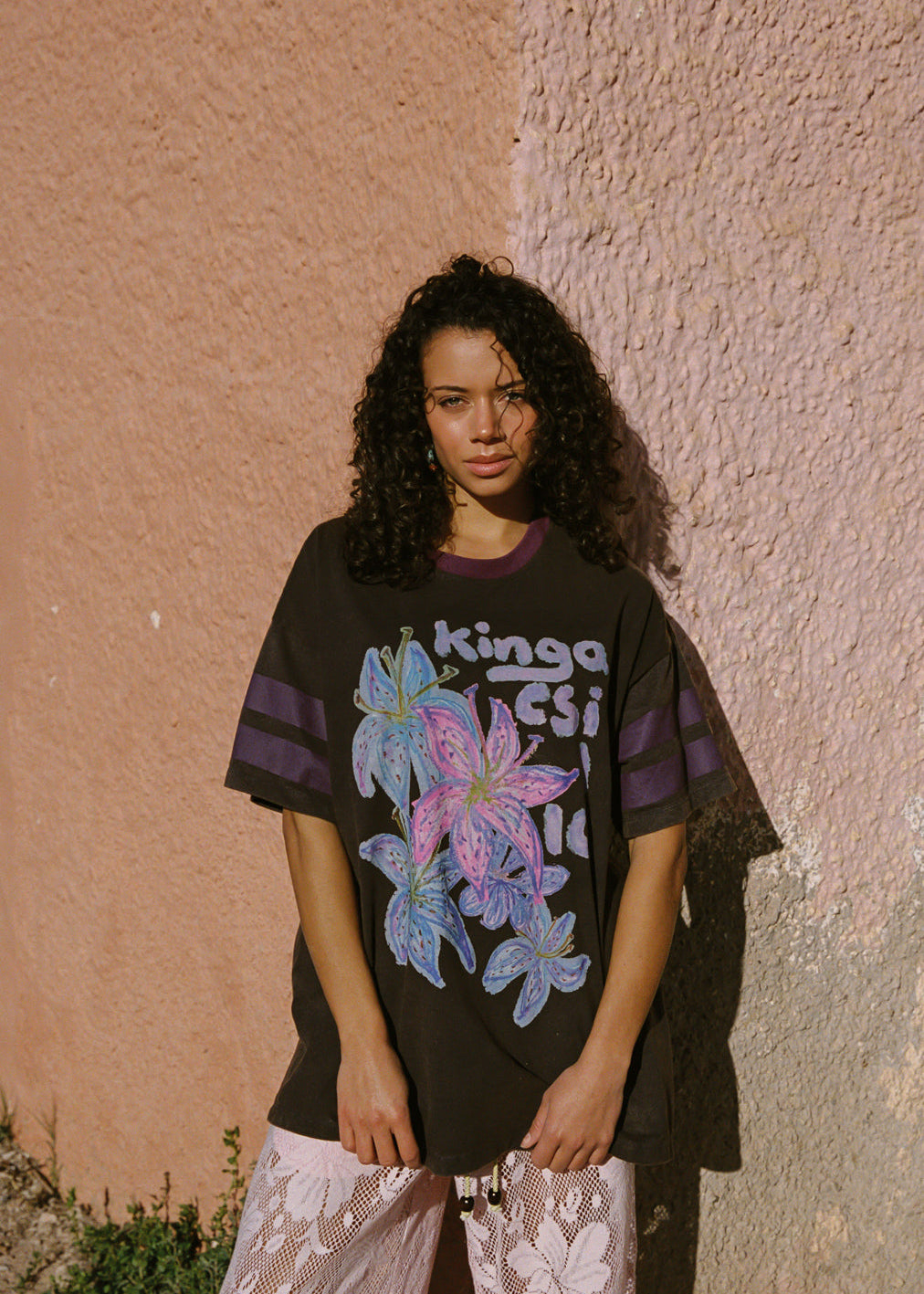 Woman wearing a black t-shirt with colorful floral design and text, standing against a textured wall.