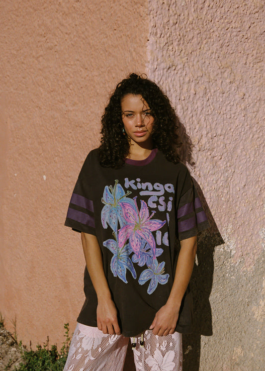 Woman wearing a black t-shirt with colorful floral design and text, standing against a textured wall.