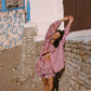 Woman in a pink patterned outfit dancing in front of a stone wall and white building.