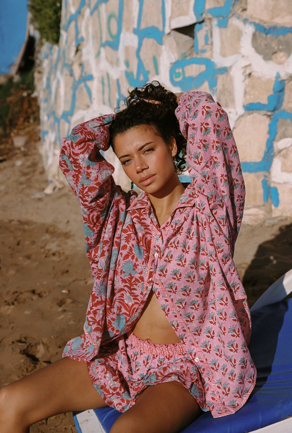 Woman in a pink floral outfit sitting against a textured wall with graffiti.