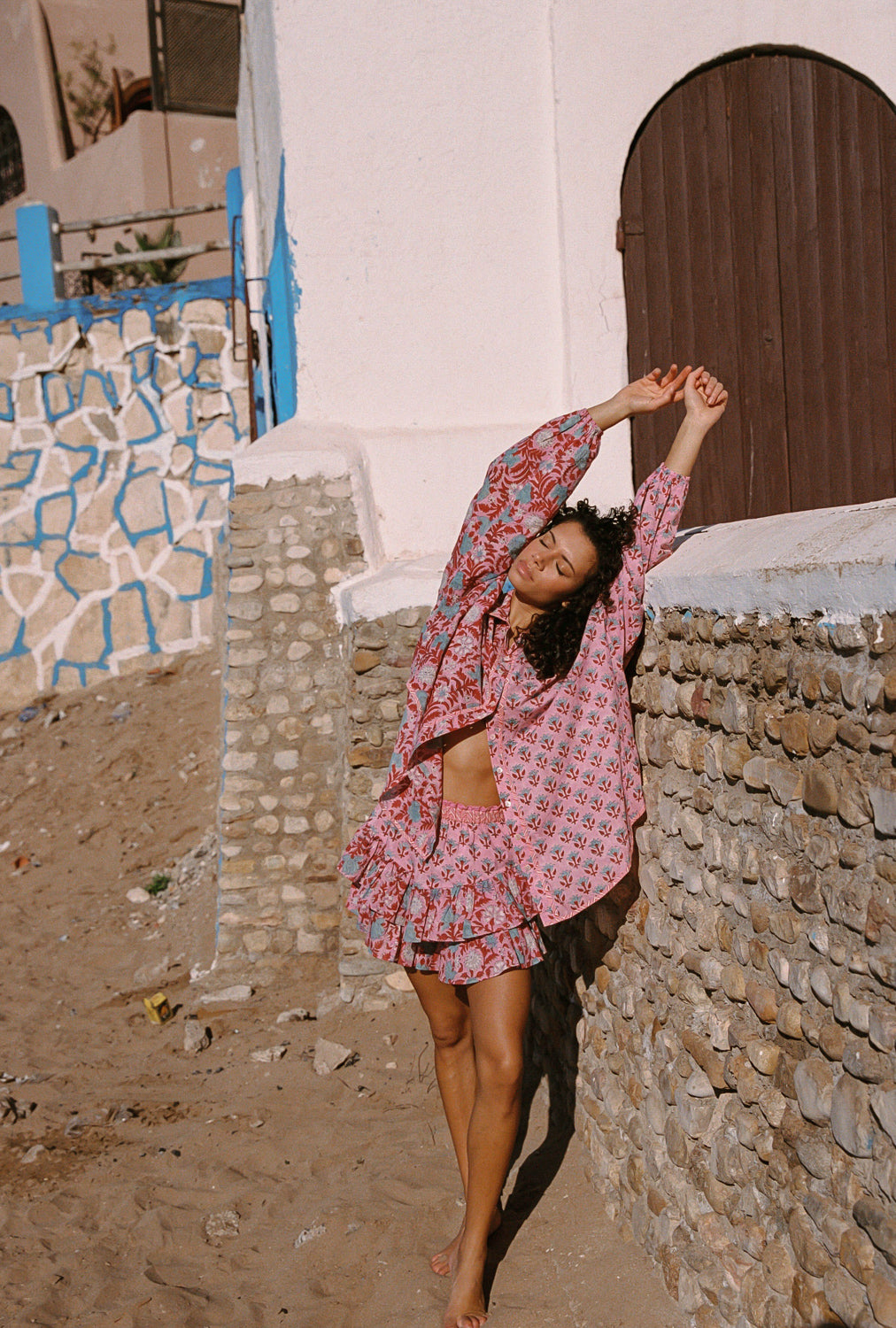 Woman in a pink patterned outfit dancing in front of a stone wall and white building.