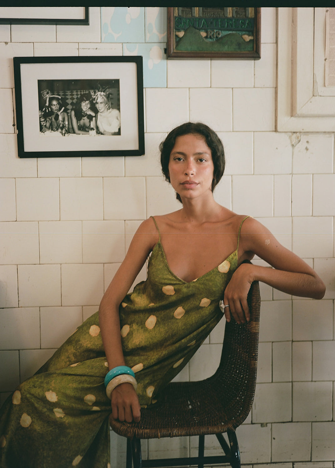 Woman in a green polka dot dress sitting on a chair against a tiled wall with framed pictures.