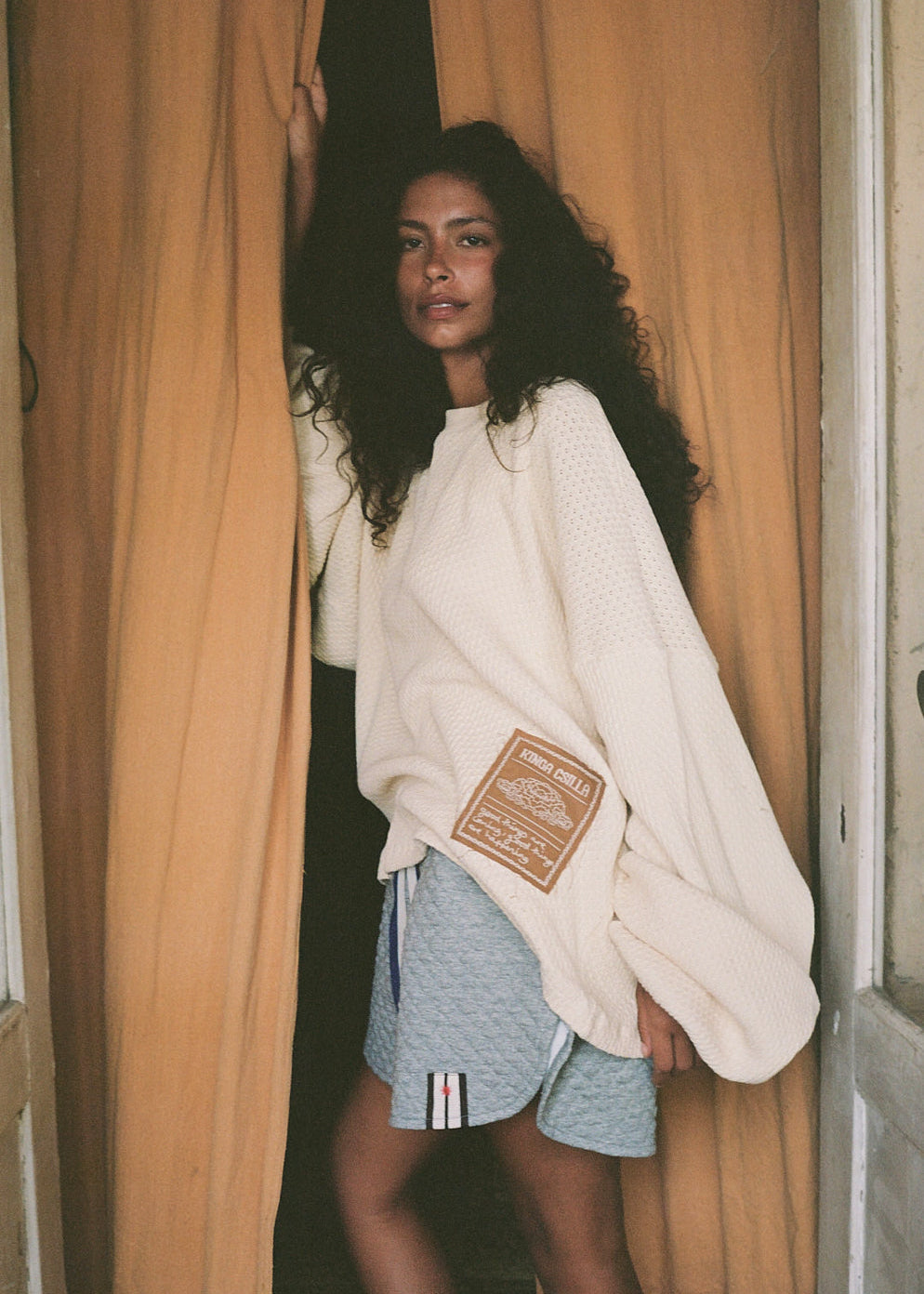 Woman wearing a white sweatshirt with a patch and blue shorts standing in front of a curtain.