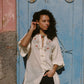 Woman wearing a white embroidered shirt and black shorts standing in front of a blue door.