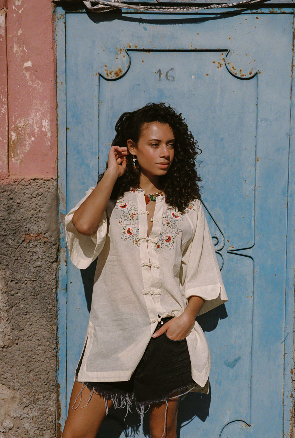 Woman wearing a white embroidered shirt and black shorts standing in front of a blue door.