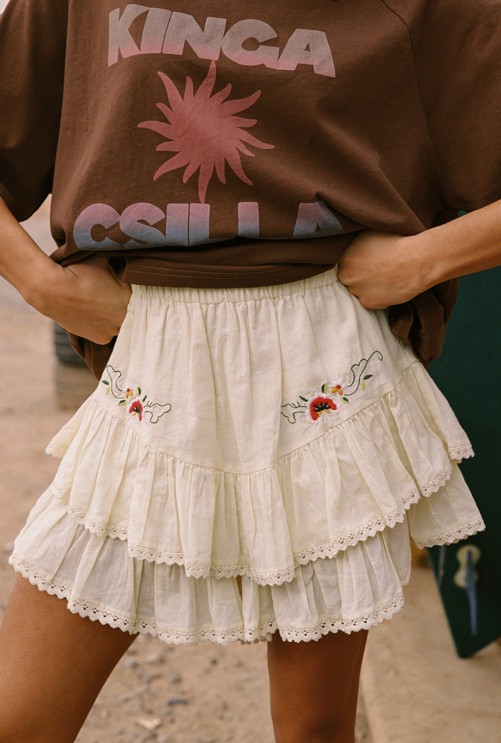 Person wearing a brown sweatshirt with pink graphics and a white embroidered skirt.