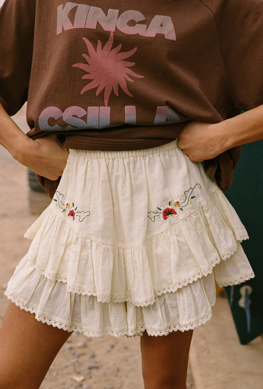Person wearing a brown sweatshirt with pink graphics and a white embroidered skirt.