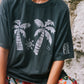 Person wearing a dark green t-shirt with palm tree design and colorful shorts sitting against a stone wall.