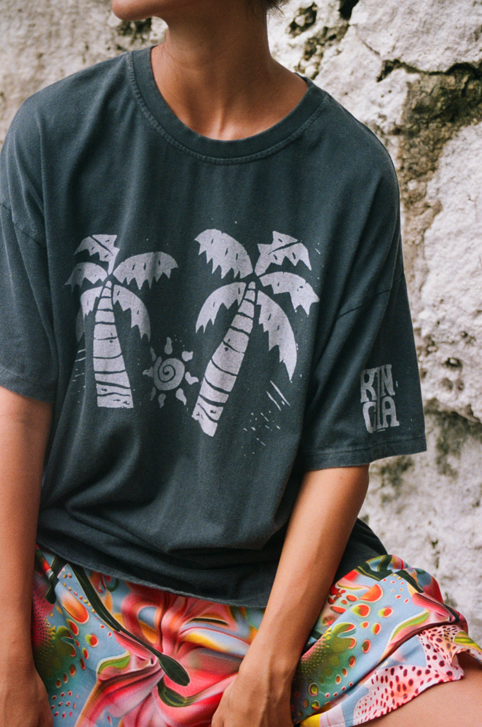 Person wearing a dark green t-shirt with palm tree design and colorful shorts sitting against a stone wall.