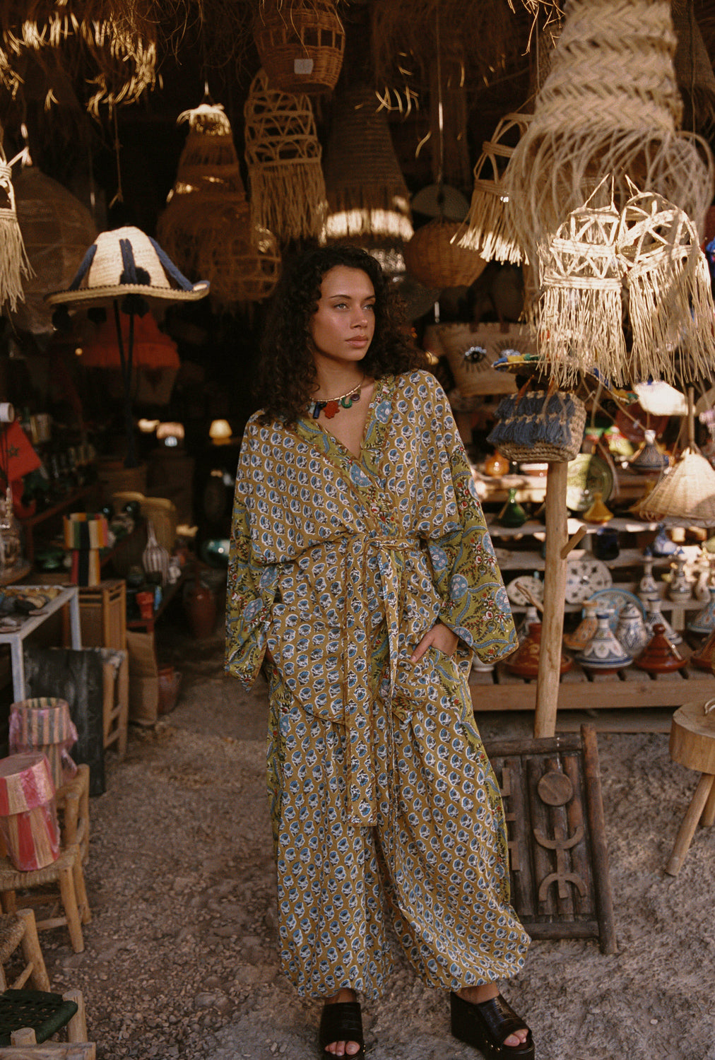 Woman in a patterned dress standing in a market with various wares hanging above her.