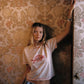 A woman stands indoors against ornate floral wallpaper, wearing a light pink cropped T-shirt with a red graphic print and blue patterned bottoms. She has bracelets on her wrist and holds a dark vertical object near the wall.
