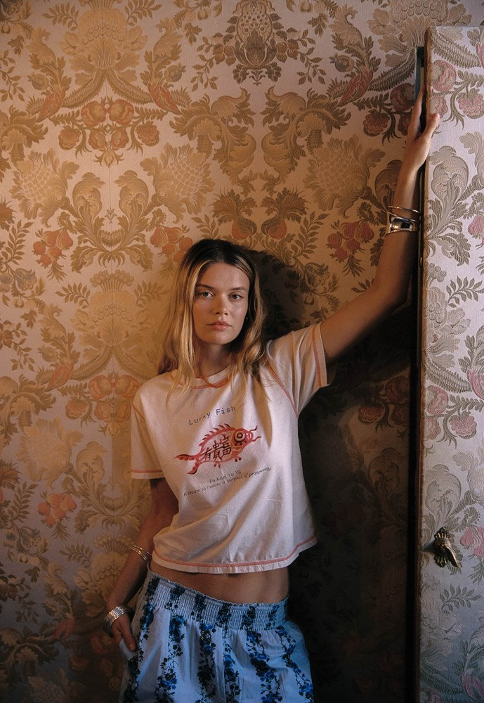 A woman stands indoors against ornate floral wallpaper, wearing a light pink cropped T-shirt with a red graphic print and blue patterned bottoms. She has bracelets on her wrist and holds a dark vertical object near the wall.