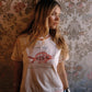 A woman with long blonde hair wearing a cream-colored graphic t-shirt with a red fish design and the text 'Lucky Fish', paired with blue patterned shorts, standing in front of a vintage floral wallpaper background.