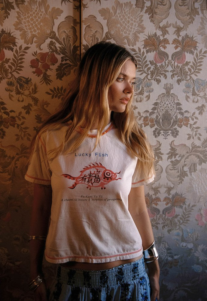 A woman with long blonde hair wearing a cream-colored graphic t-shirt with a red fish design and the text 'Lucky Fish', paired with blue patterned shorts, standing in front of a vintage floral wallpaper background.