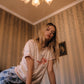 A woman with long blonde hair wearing a cream-colored graphic t-shirt with a red fish design and the text 'Lucky Fish', paired with blue patterned shorts.