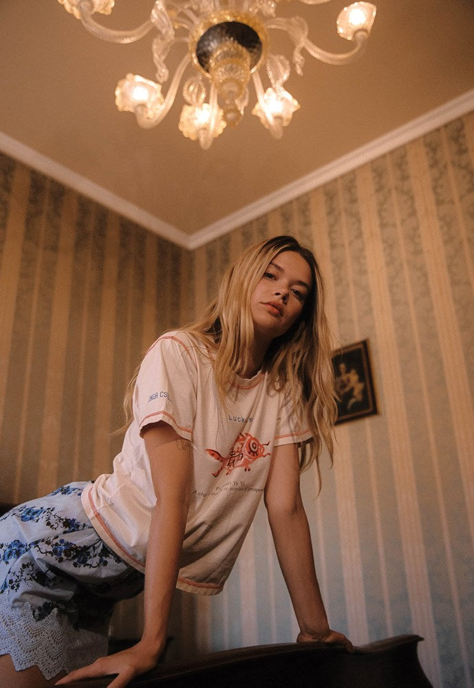 A woman with long blonde hair wearing a cream-colored graphic t-shirt with a red fish design and the text 'Lucky Fish', paired with blue patterned shorts.