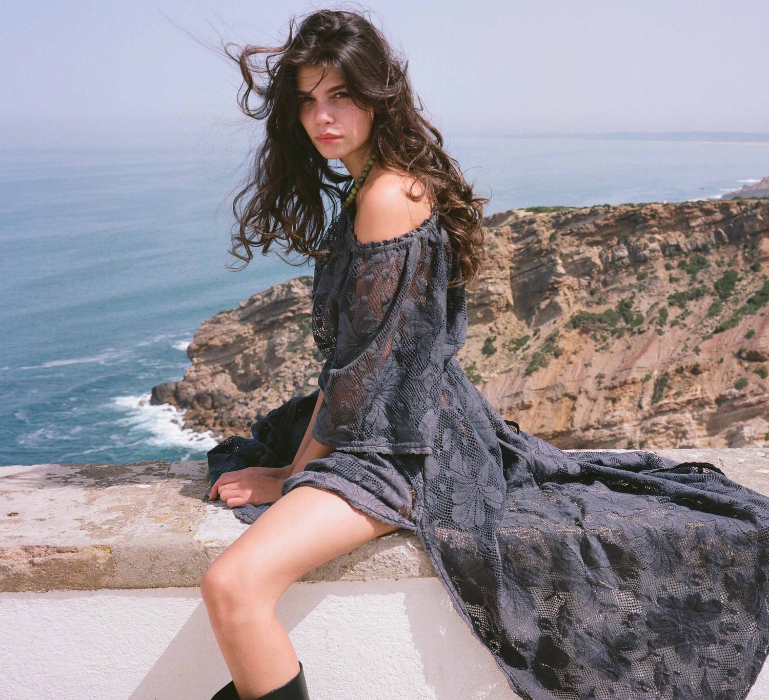 Woman in a dark gray dress sitting on a ledge with ocean and cliffs in the background