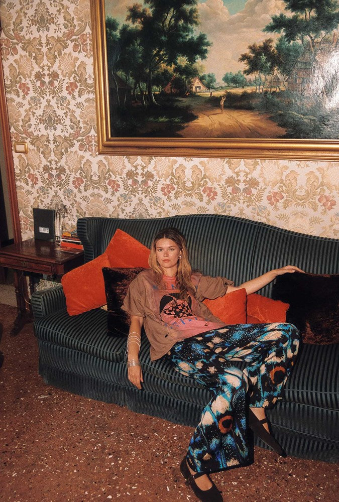 A woman sits on a dark green ribbed sofa in a vintage room with patterned wallpaper and a large framed landscape painting. She is wearing a brown graphic t-shirt, a pink top, and blue, black, and orange patterned trousers.
