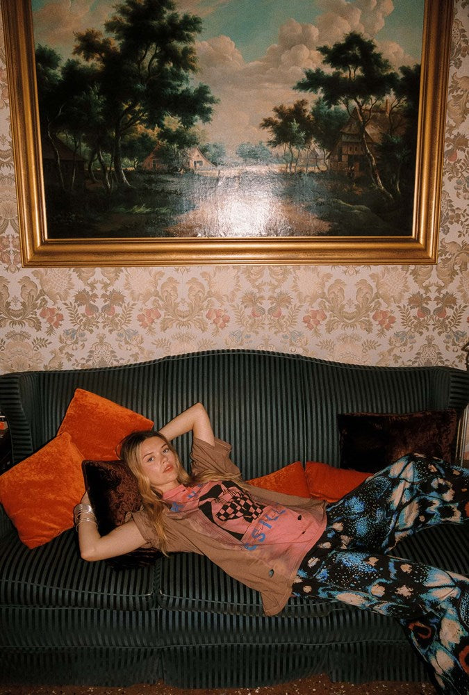 A woman reclines on a dark green ribbed sofa in a vintage room, wearing a pink graphic t-shirt and blue-and-white patterned trousers. A large framed landscape painting hangs on the patterned wallpapered wall above the couch.