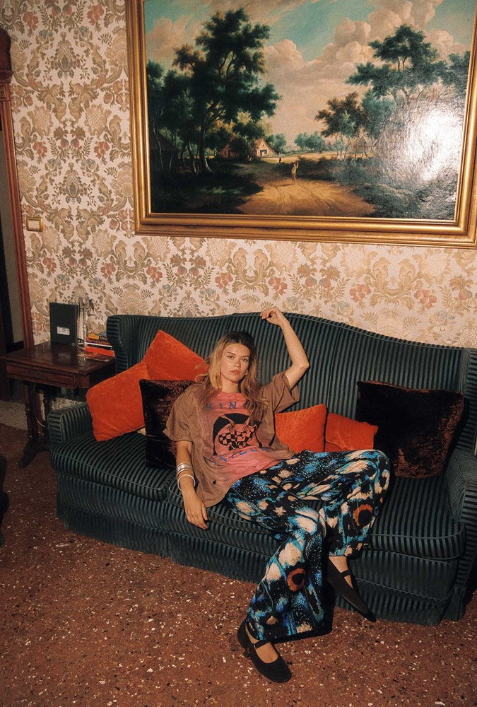 A woman sits on a dark green striped sofa in a vintage room with patterned wallpaper and a large landscape painting on the wall. She is wearing a brown and pink graphic t-shirt, a pink top, and blue floral print pants.