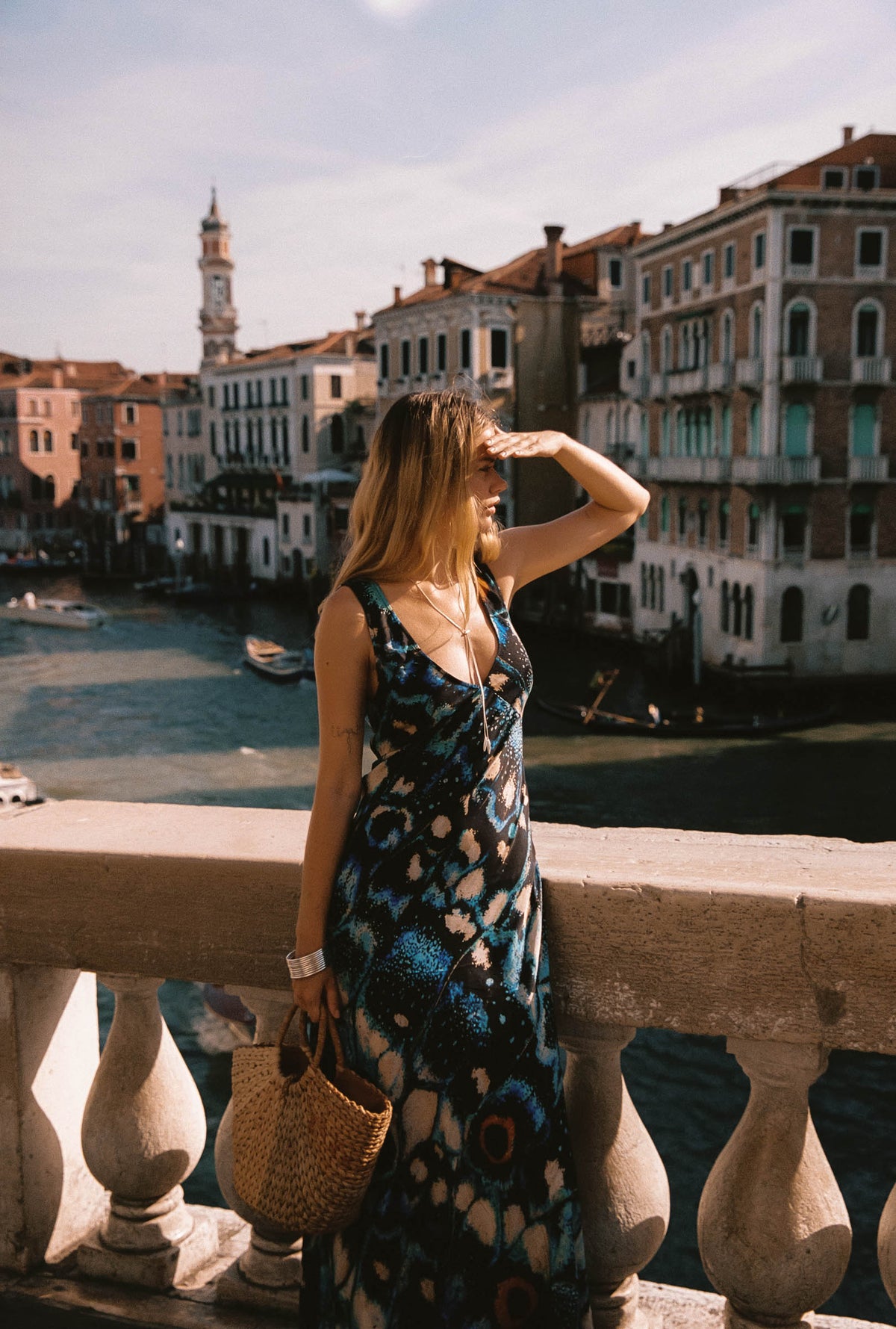 A woman wearing a sleeveless blue and black patterned maxi dress stands on a stone balcony overlooking a canal with historic buildings in Venice, holding a woven straw bag.