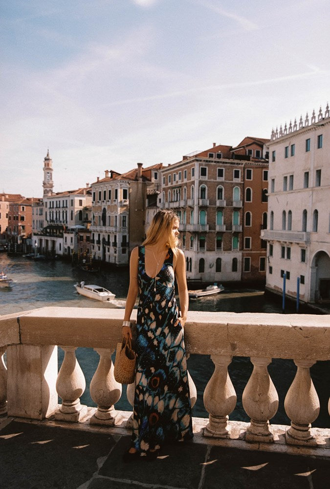 A woman wearing a sleeveless blue and black patterned maxi dress stands on a stone balcony overlooking a canal with historic buildings in Venice, holding a woven straw bag.