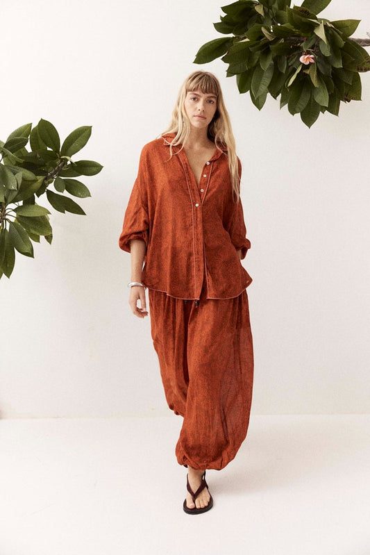 A woman modeling a rust-orange two-piece linen pajama set consisting of a button-down shirt and matching harem pants, styled with black flip-flops against a white studio background with green foliage props.