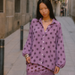 A woman stands on a narrow city street wearing a lavender-purple long-sleeve button-up shirt and matching skirt set with dark brown polka dots and a scalloped lace hem.