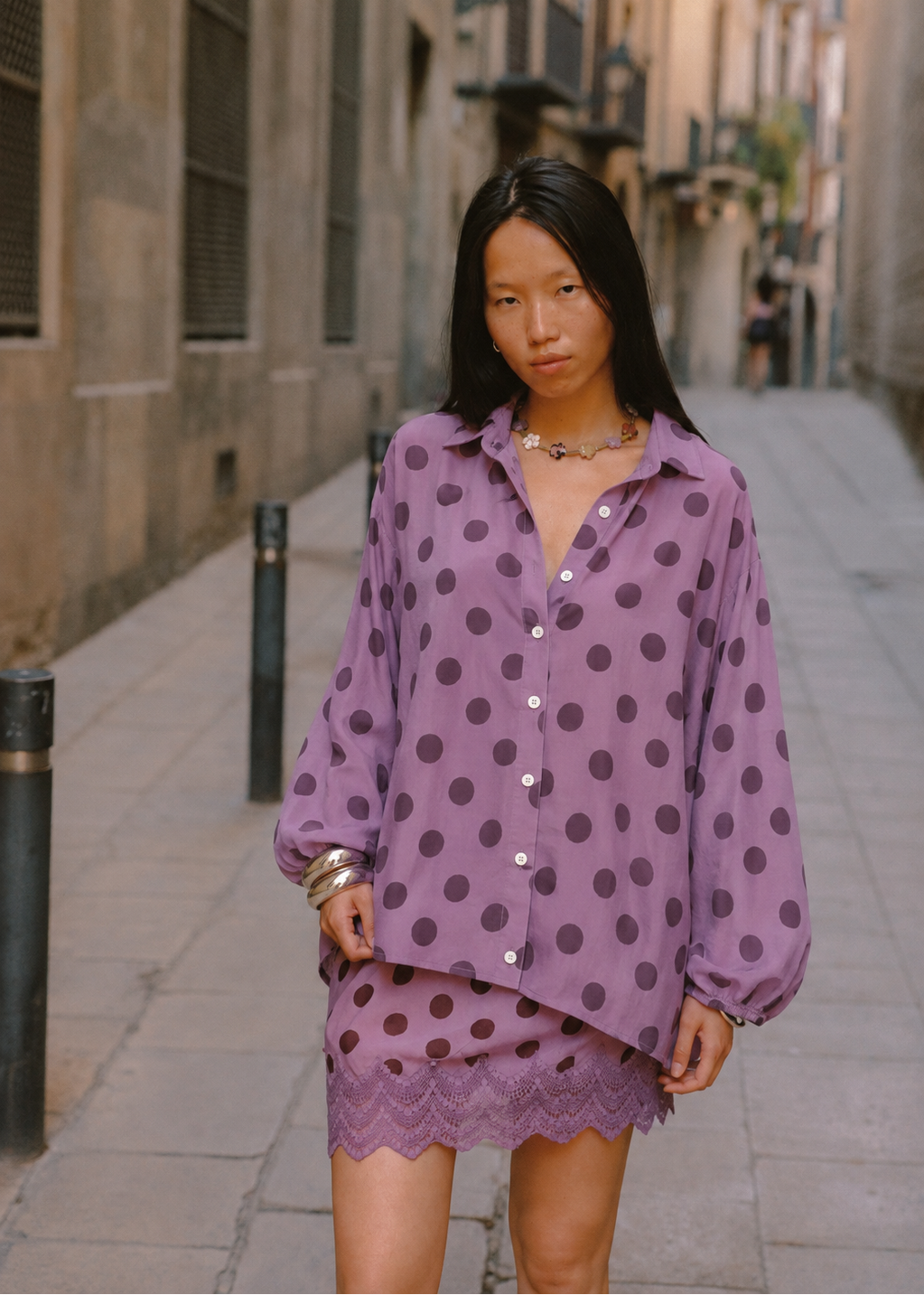 A woman stands on a narrow city street wearing a lavender-purple long-sleeve button-up shirt and matching skirt set with dark brown polka dots and a scalloped lace hem.
