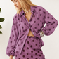 A woman wearing a matching purple and dark brown polka-dot two-piece set, featuring a long-sleeve button-up shirt and a high-waisted mini skirt with a lace hem, set against a white wall with green leaves.