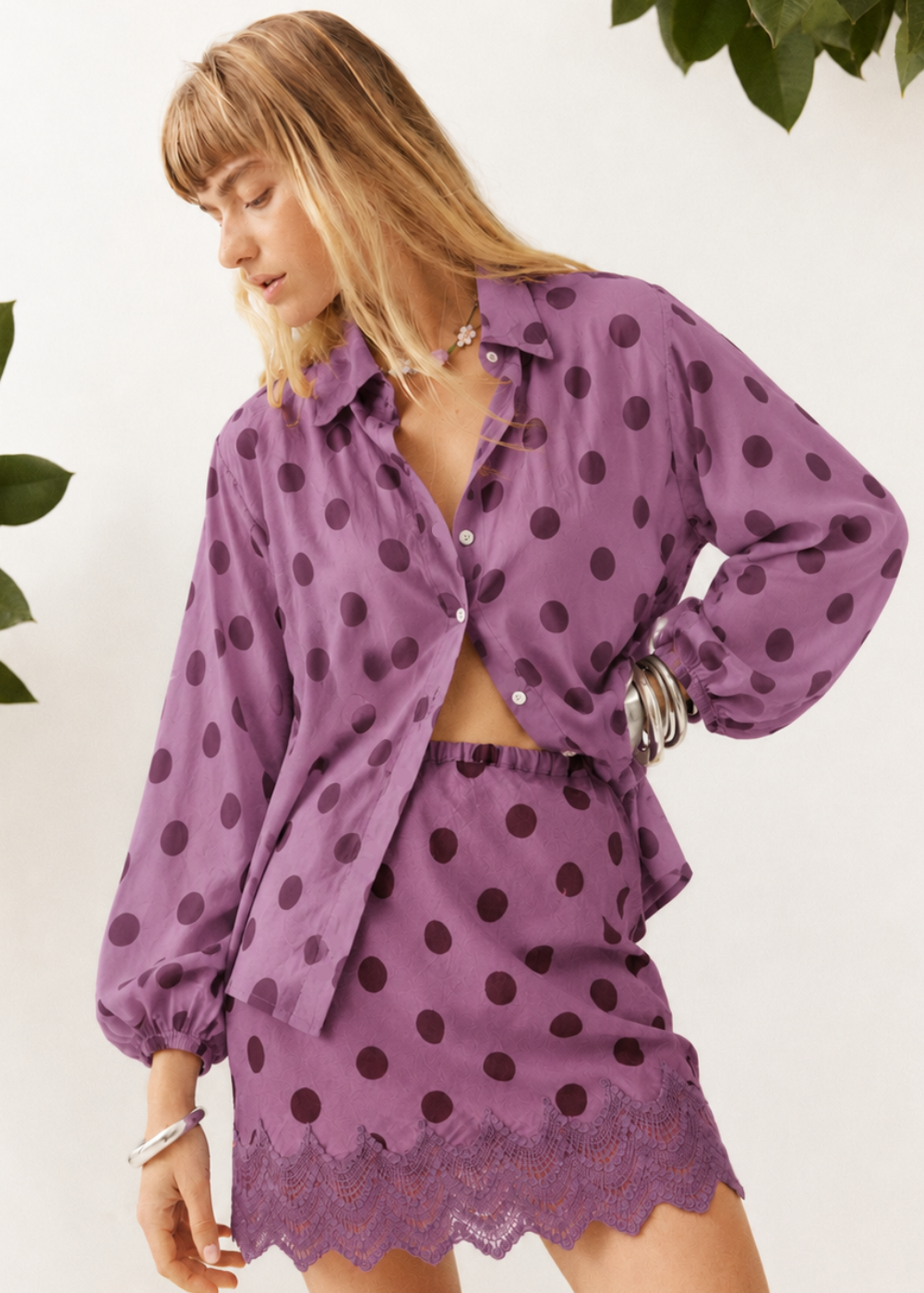 A woman wearing a matching purple and dark brown polka-dot two-piece set, featuring a long-sleeve button-up shirt and a high-waisted mini skirt with a lace hem, set against a white wall with green leaves.