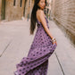 A woman wearing a flowing lavender-purple maxi dress with black polka dots stands on a stone sidewalk in a narrow European-style street, holding out the side of the dress to show its movement.