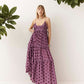 A woman wearing a purple tiered maxi dress with dark brown polka dots, standing against a white wall with green plants in the corners.