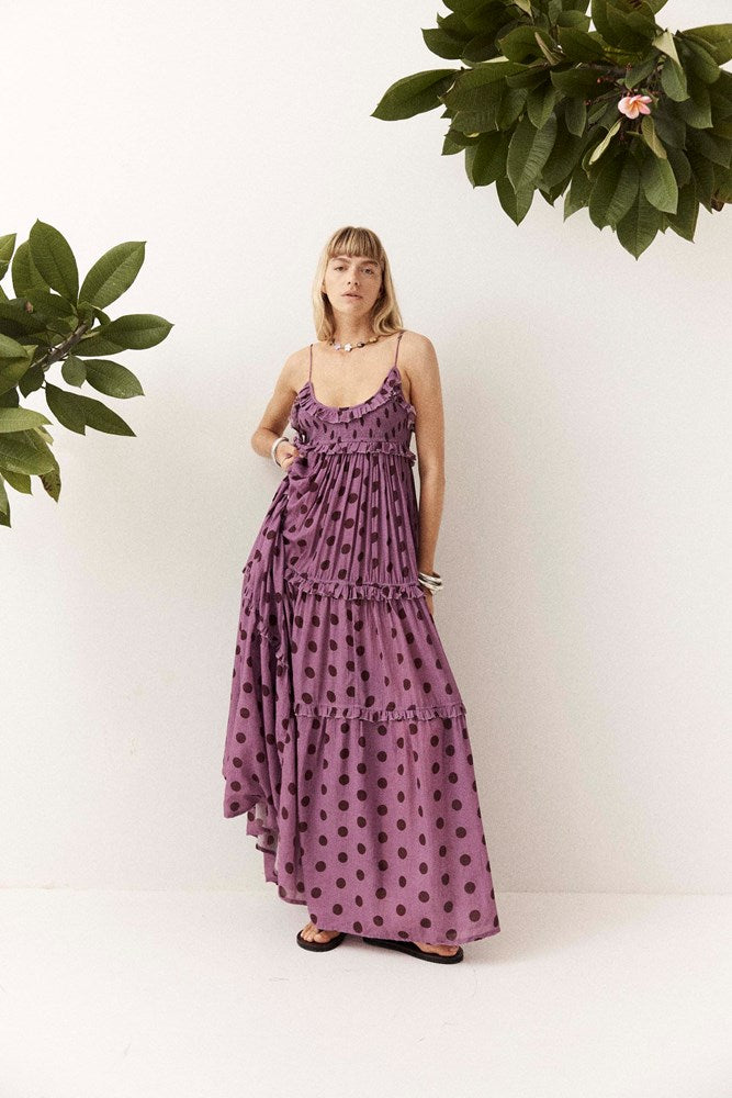 A woman wearing a purple tiered maxi dress with dark brown polka dots, standing against a white wall with green plants in the corners.