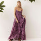 A woman wearing a purple tiered maxi dress with dark brown polka dots, standing against a white wall with green plants in the corners.