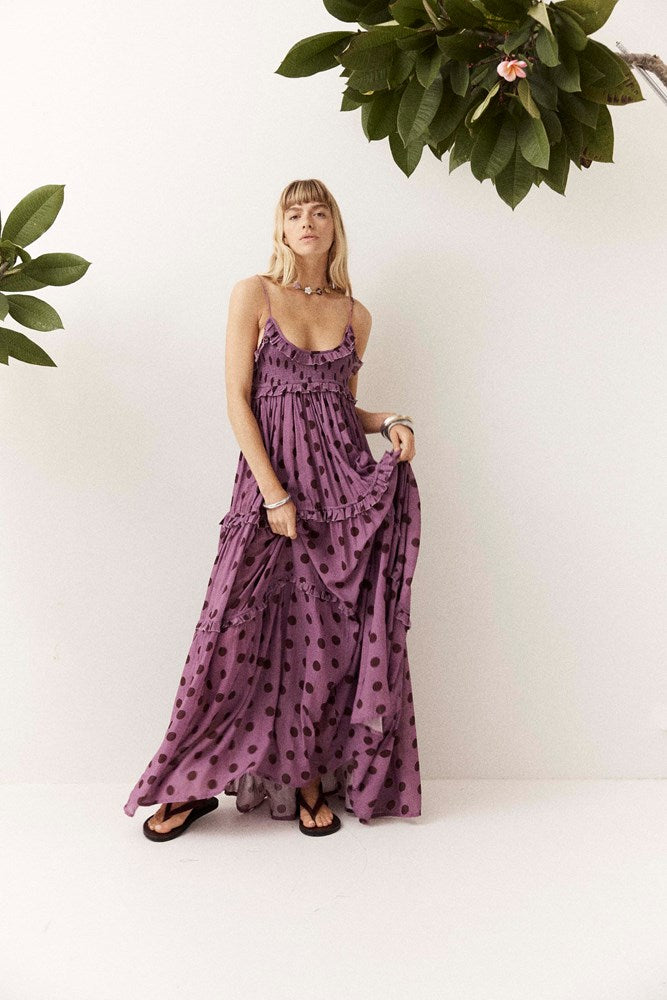 A woman wearing a purple tiered maxi dress with dark brown polka dots, standing against a white wall with green plants in the corners.