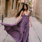 A woman wearing a purple tiered maxi dress with dark brown polka dots walks down a narrow stone street, holding out the side of the dress to show its flow. She is wearing silver bracelets and black flat shoes.