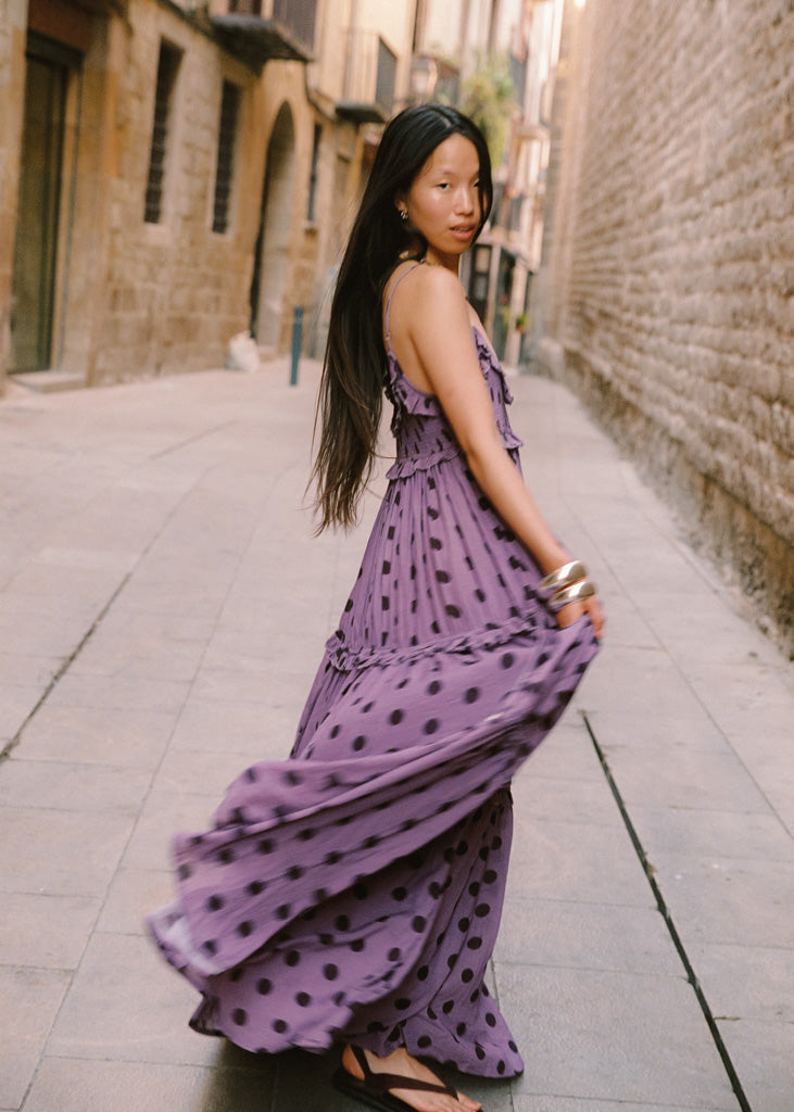 A woman wearing a flowing lavender-purple maxi dress with black polka dots stands on a stone sidewalk in a narrow European-style street, holding out the side of the dress to show its movement.
