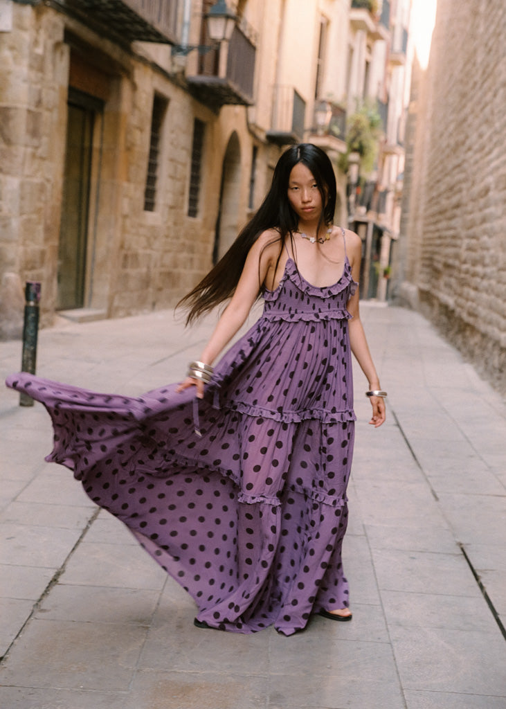 A woman wearing a purple tiered maxi dress with dark brown polka dots walks down a narrow stone street, holding out the side of the dress to show its flow. She is wearing silver bracelets and black flat shoes.