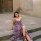 A woman sits on stone steps wearing a purple sleeveless maxi dress with a dark brown polka-dot pattern and dark brown strappy sandals, set against a stone wall and a large wooden door.