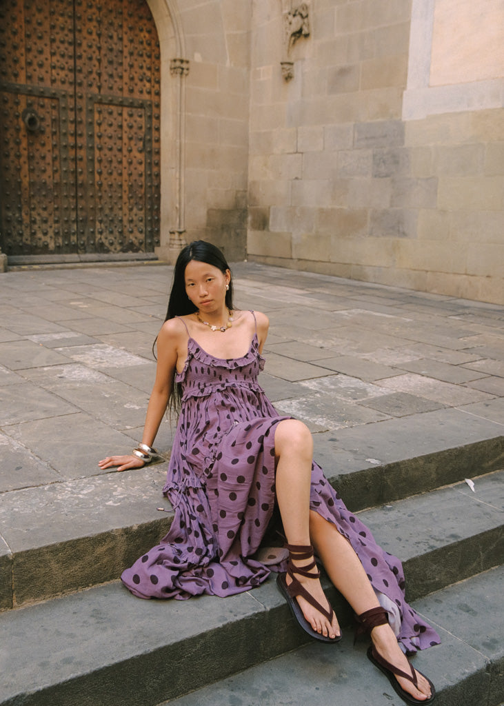 A woman sits on stone steps wearing a purple sleeveless maxi dress with a dark brown polka-dot pattern and dark brown strappy sandals, set against a stone wall and a large wooden door.