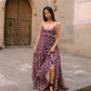 A woman wearing a purple maxi dress with dark brown polka dots stands on stone steps in front of a stone building with a large wooden door.